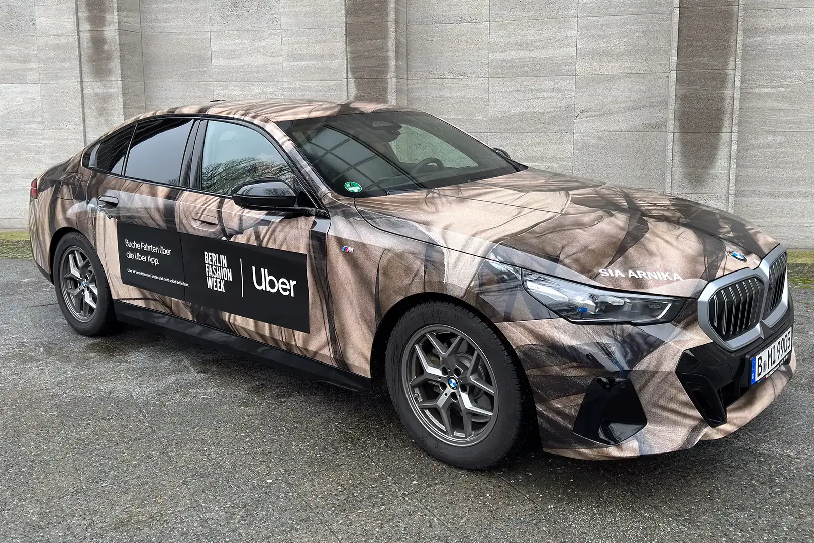 Ein moderner BMW, foliert im abstrakten, braun-schwarzen Stoffmuster der Designerin Sia Arnika. Auf der Motorhaube steht ‚SIA ARNIKA‘, auf der Seite befinden sich Logos von Uber, Berlin Fashion Week und der Slogan ‚Buche Fahrten über die Uber App‘